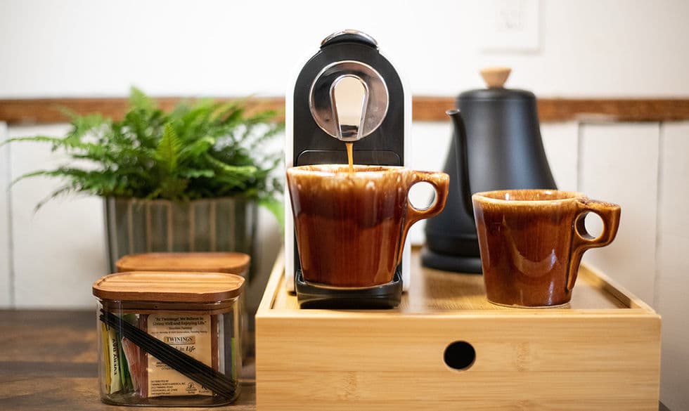 Coffee bar with coffee maker, brown pottery mugs, an electric tea kettle, and glass containers with wooden tops holding an assortment of teas.