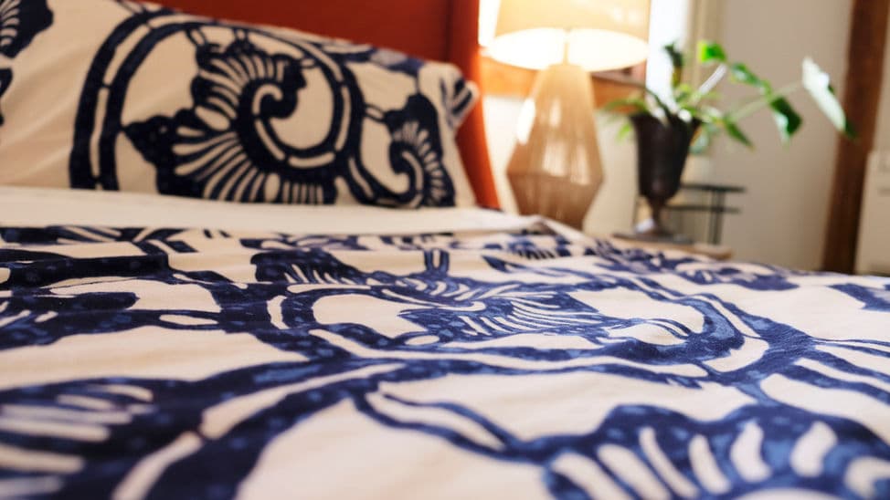 Close up of a king bed with a blue and cream patterned bedspread and matching pillows and a table lamp nearby.