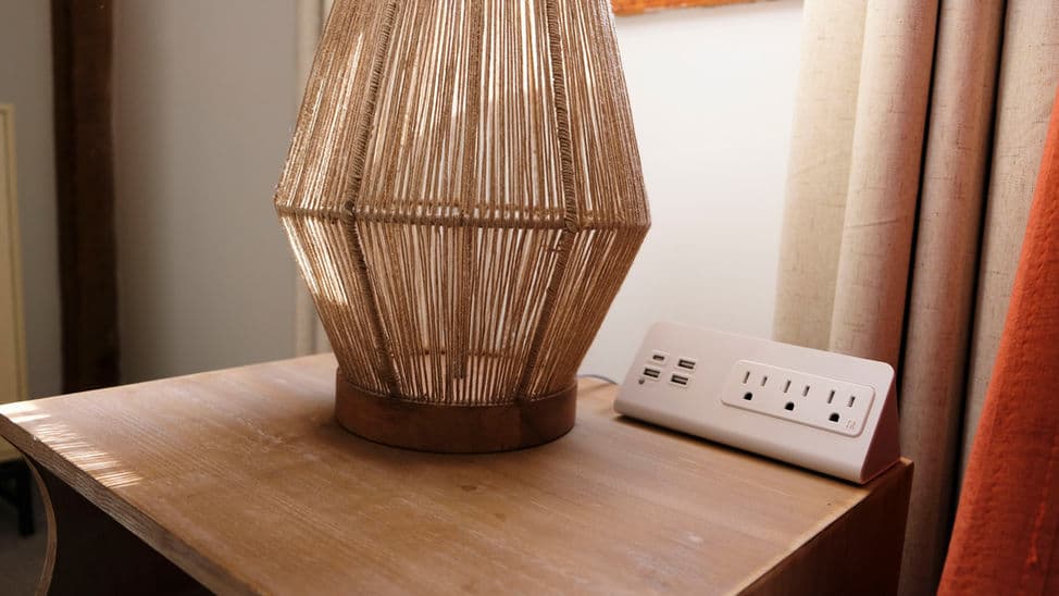 Close up of the top of a nightstand with natural fiber lamp base and power strip with three outlets and four charging ports.