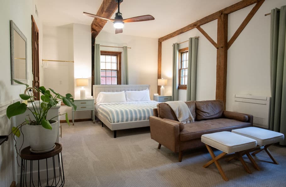 Tranquil bedroom with king bed with loveseat, ceiling fan, exposed beams, and natural light from several windows.
