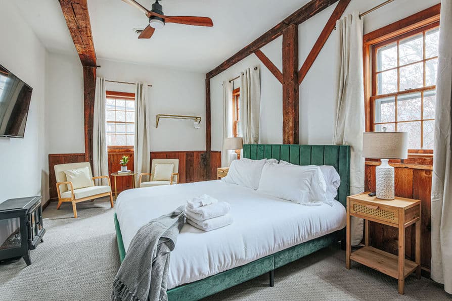 Light and airy bedroom with king bed, matching nightstands with table lamps, exposed beams, and wall-mounted smart TV.