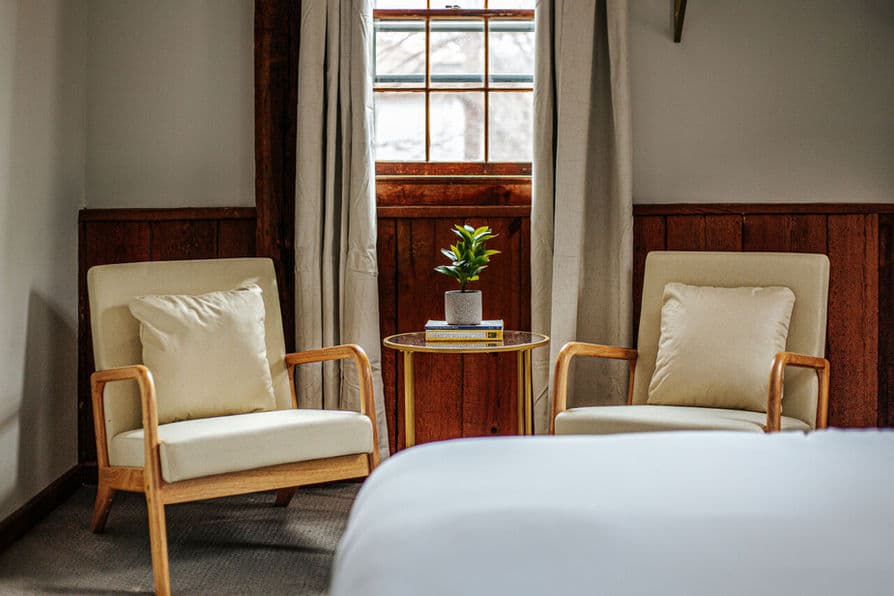 Two wooden armchairs with cream cushions flank a small, round occasional table under a window in the bedroom.