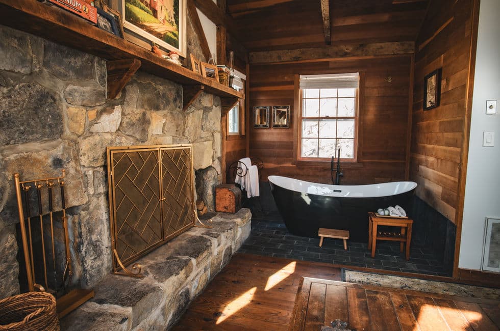 Rustic cabin interior with large stone fireplace, wood paneled walls, and black & white slipper tub.