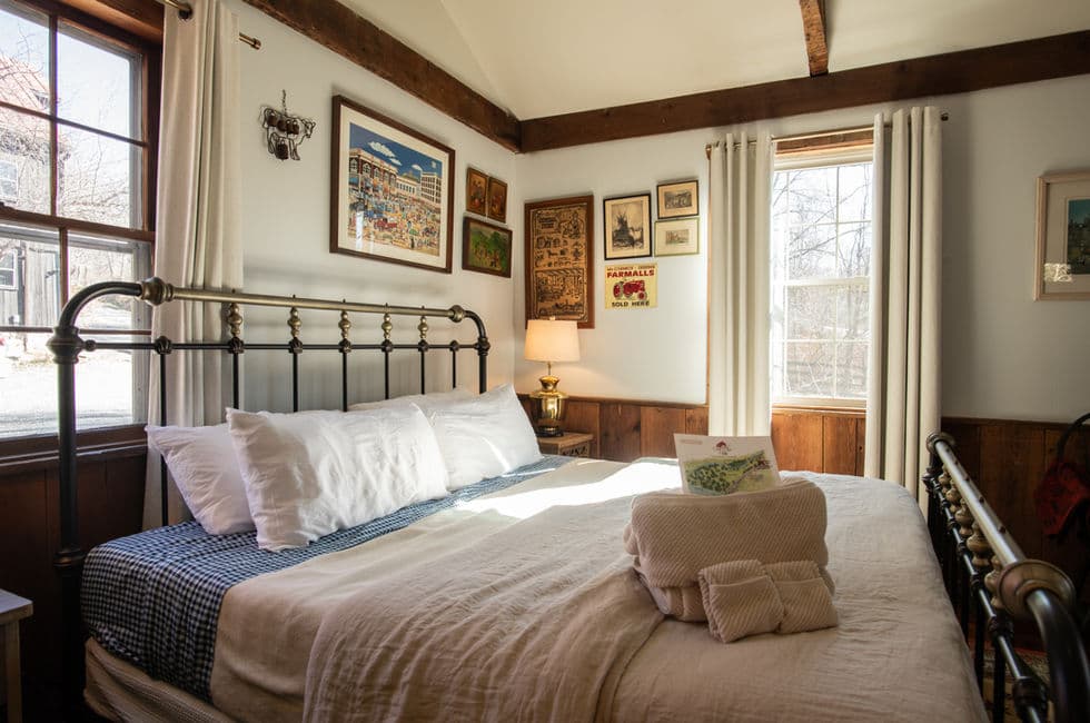 A bedroom with metal frame king-size bed with loft bedding and neatly stacked towels at the ready. Sunlight comes in the windows, and there is an intriguing arrangement of artwork on the walls.