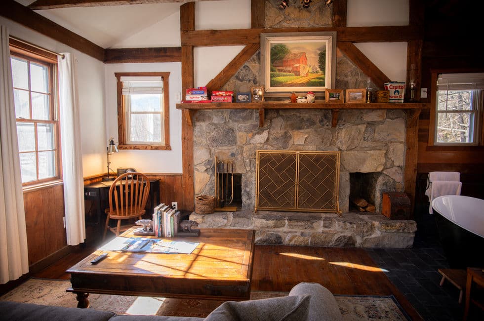 Opposite a cozy loveseat and wooden coffee table is a large stone fireplace, and to the right of the fireplace is a black & white slipper tub.