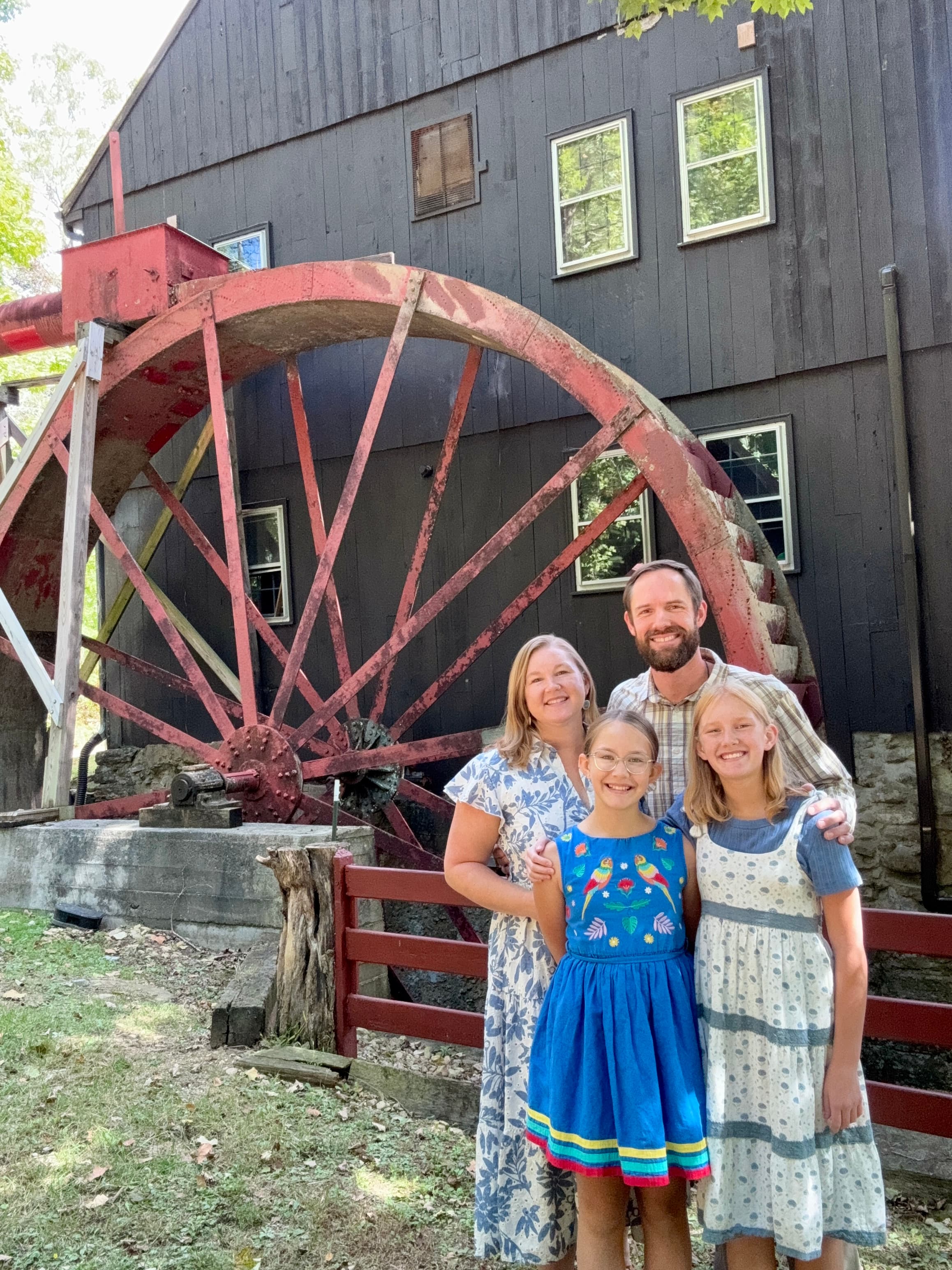 A family of four poses in front of a large, red waterwheel and a rustic building.
