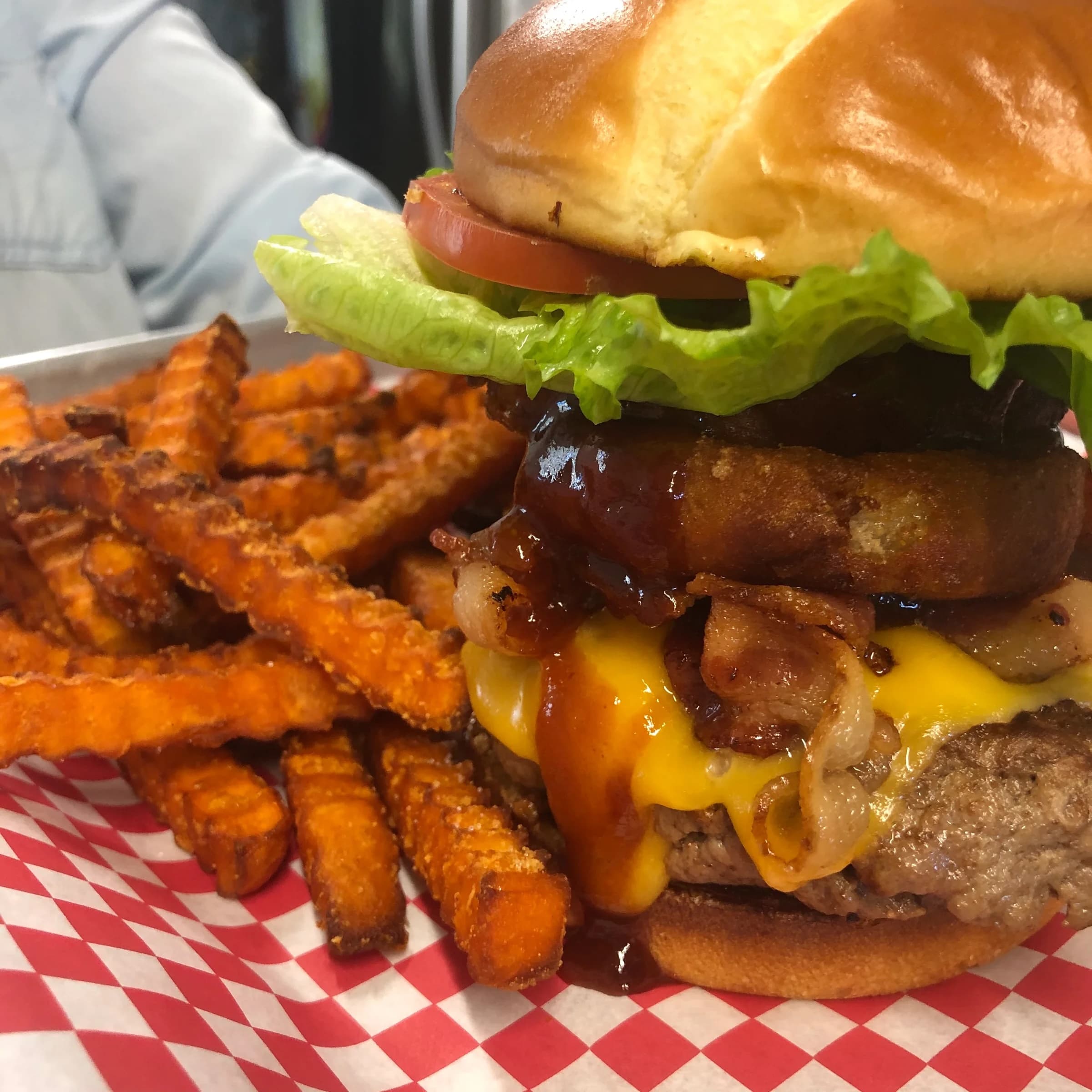 A stacked hamburger with lettuce, tomato, bacon, and cheese, served alongside sweet potato fries.