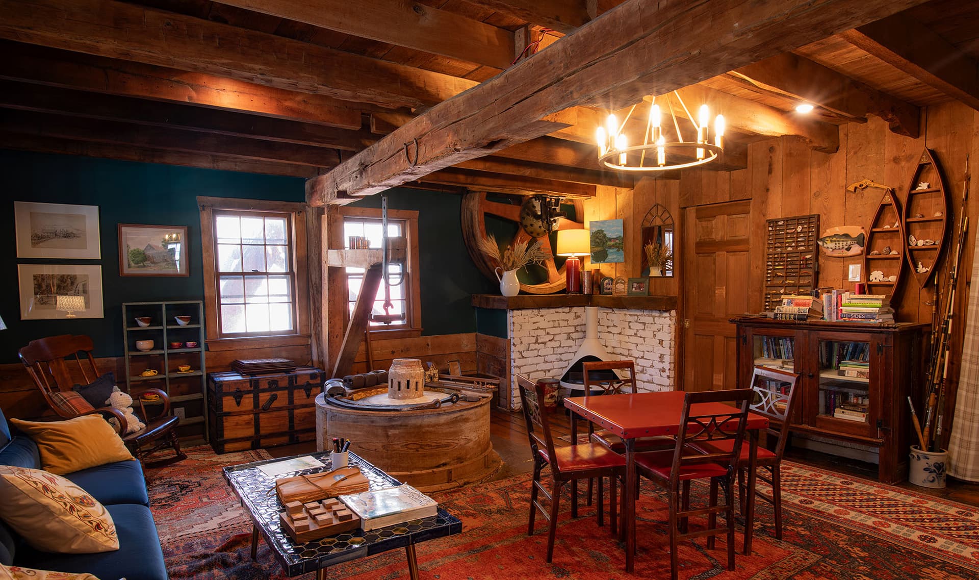 A cozy, rustic living room with wooden beams, a red dining table, and a stone fireplace, surrounded by warm décor.