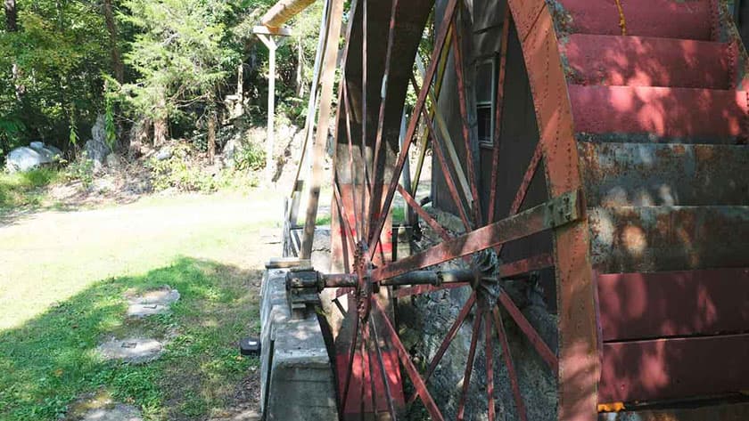 Close-up of an old waterwheel with a rustic red finish, set in a grassy area surrounded by trees.