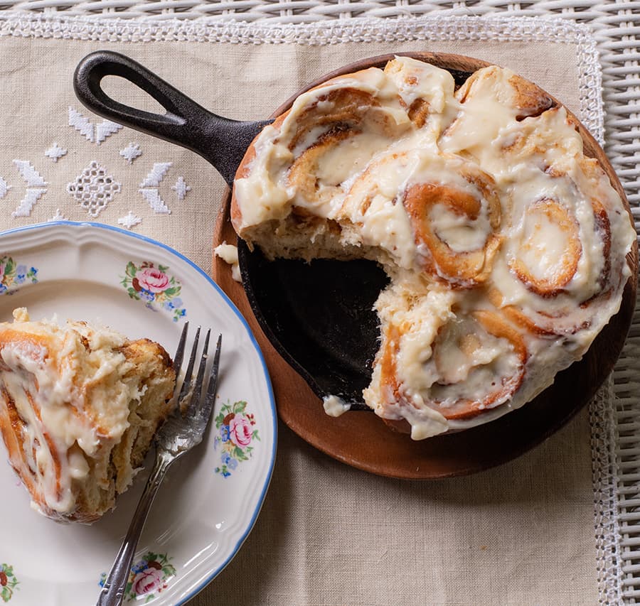 A freshly baked cinnamon roll with cream frosting on a wooden plate alongside a served slice on a floral-patterned dish with a fork.