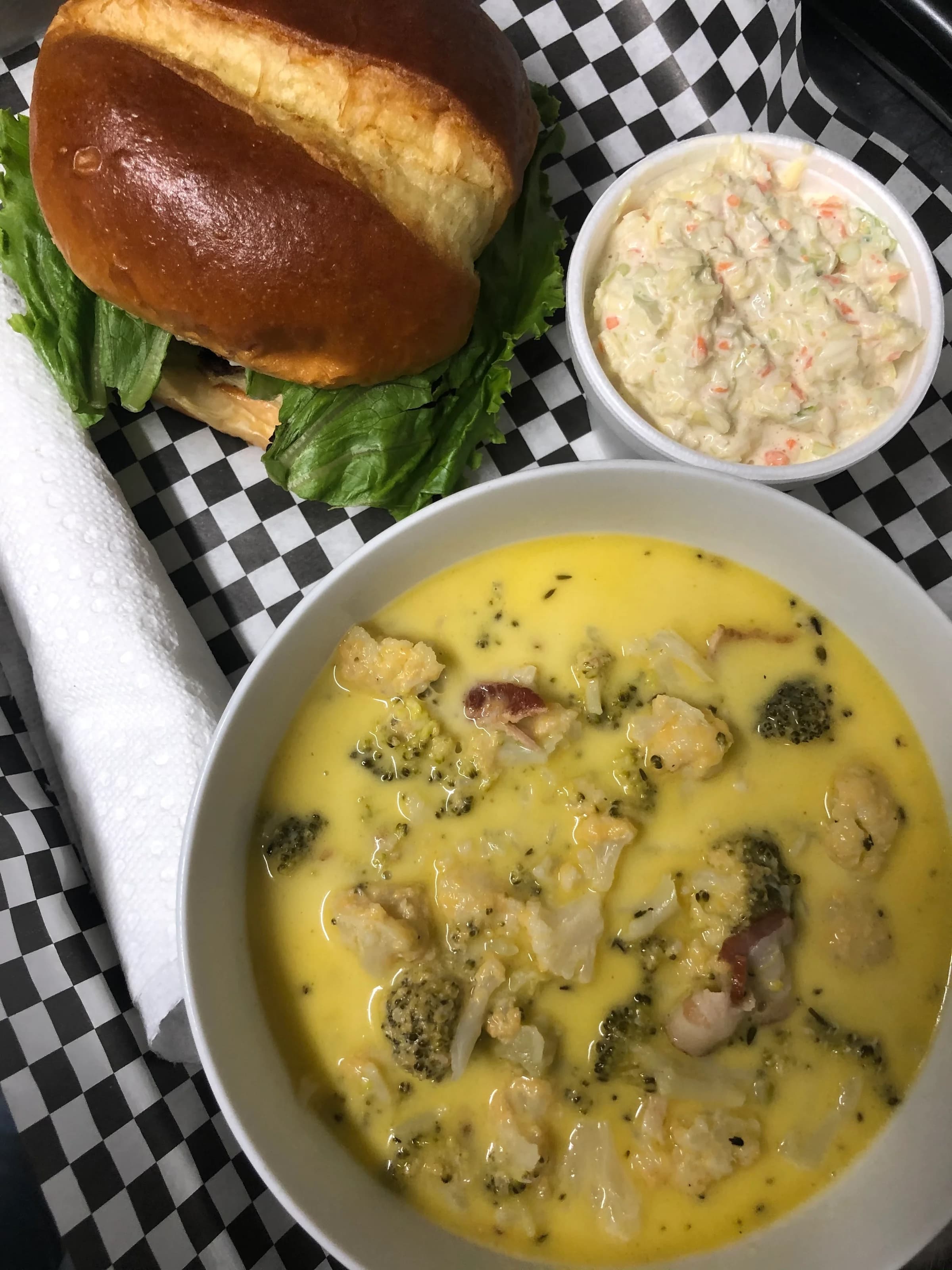 A bowl of creamy broccoli cheese soup, accompanied by a sandwich on a brioche bun and a side of coleslaw.