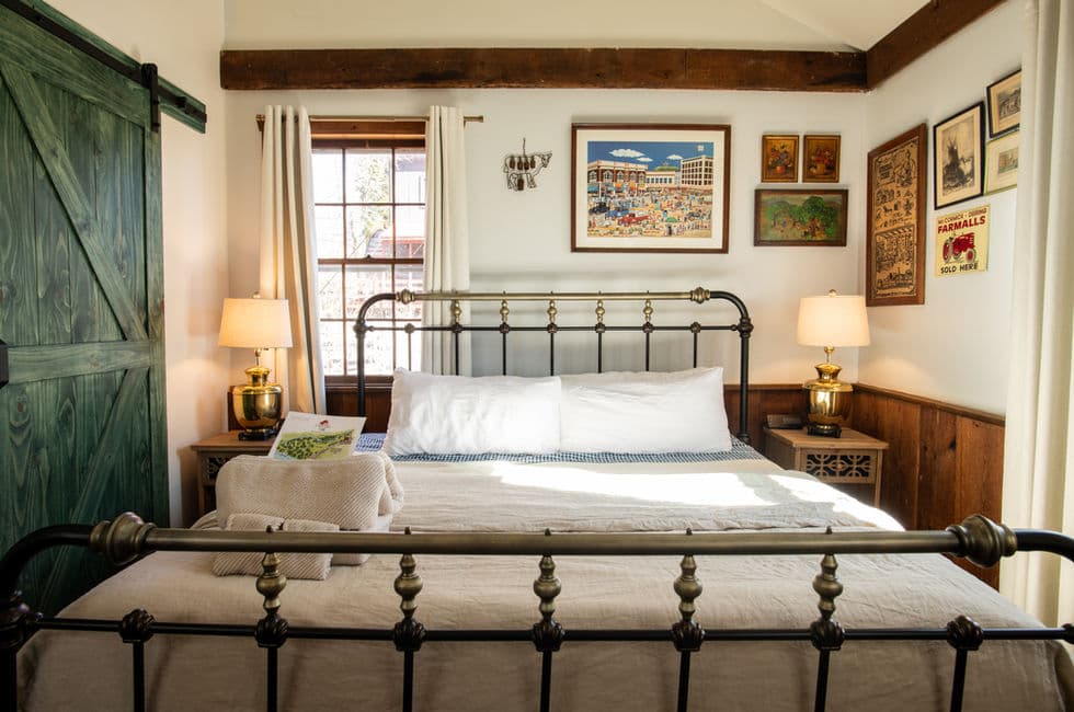 A king-size metal framed bed flanked by wooden nightstands topped with table lamps. There's a green barn style sliding door to the left and an interesting arrangement of artwork on the walls.