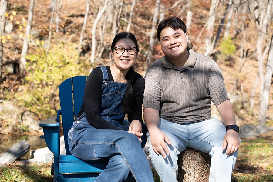 A woman and a young man sit together on a log in a wooded area, smiling at the camera.