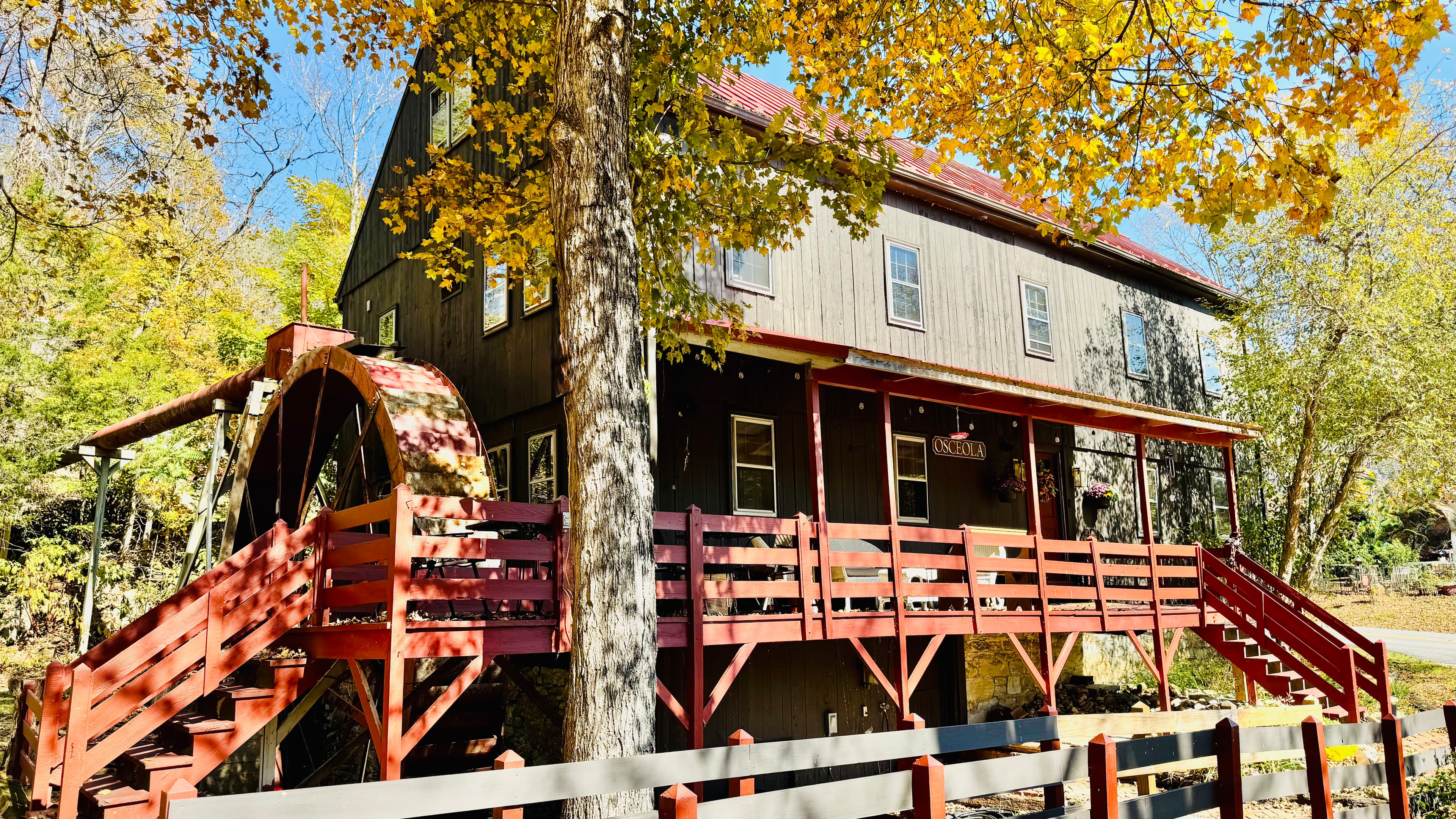 A rustic wooden building with a red water wheel and a staircase surrounded by autumn foliage.