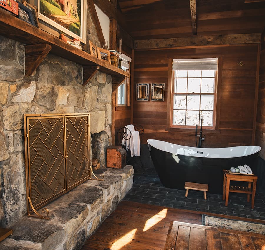 A rustic bathroom featuring a black freestanding bathtub, stone fireplace, wooden walls, and natural light from a window.